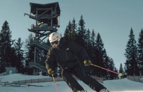 Skifahrer in schwarzer Kleidung fährt auf präparierter Piste am Semmering Hirschenkogel, im Hintergrund ein Aussichtsturm und Bäume.