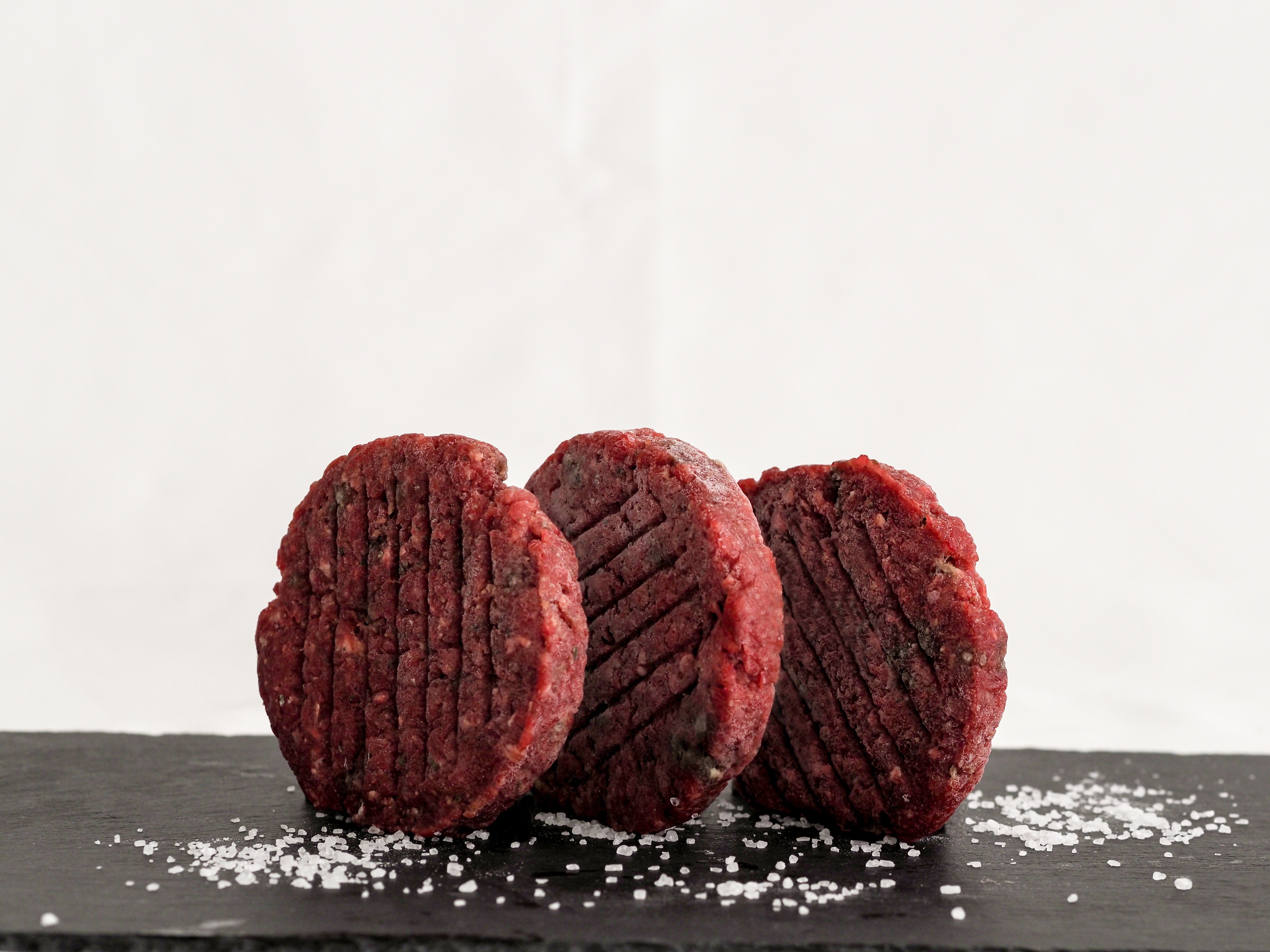 Drei rohe Burgerpatties befinden sich stehend auf einer dunklen Schieferplatte, daneben ist Salz.