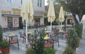 Ein leerer Schanigarten mit Tischen und Stühlen vor einem Café mit gelben Sonnenschirmen.