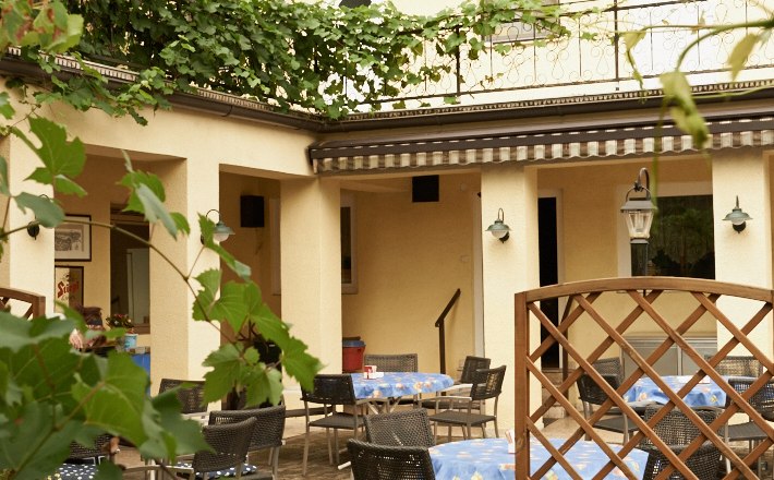 Ein gem&uuml;tlicher Gastgarten mit Tischen und St&uuml;hlen, umgeben von gr&uuml;nen Bl&auml;ttern und einem gelben Geb&auml;ude im Hintergrund.