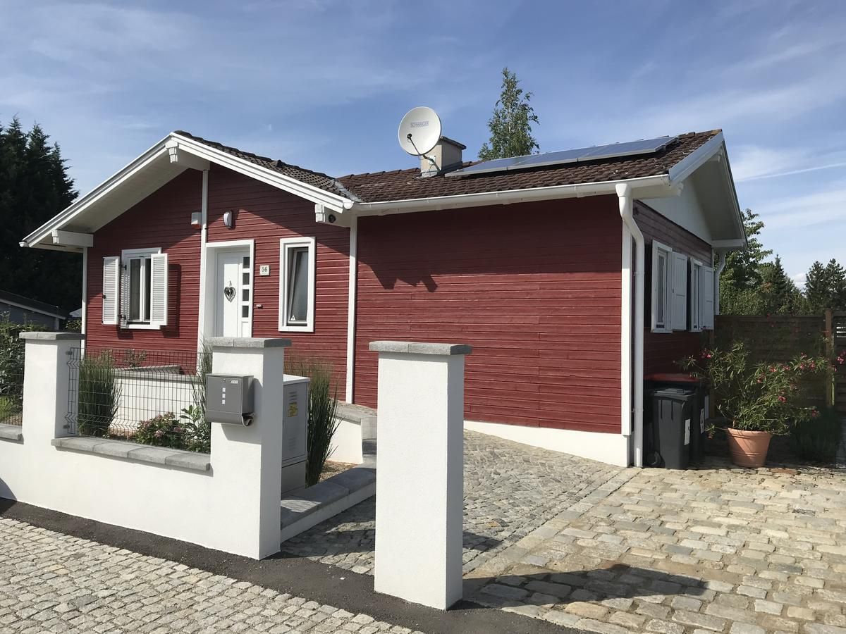 Rotes Holzhaus mit weißen Fensterläden und Solarpanelen auf dem Dach, umgeben von einem gepflasterten Hof und einer niedrigen Mauer.