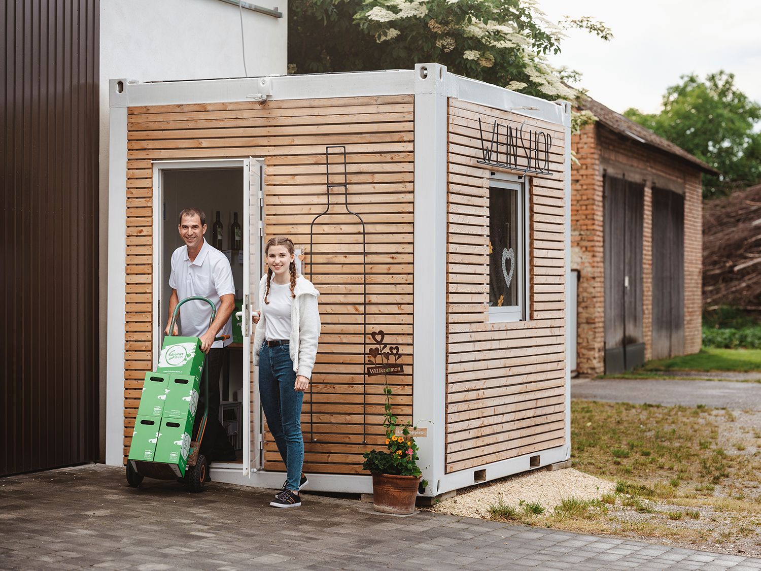 Zwei Personen vor einem kleinen Holzgebäude mit der Aufschrift 'Weinshop'.