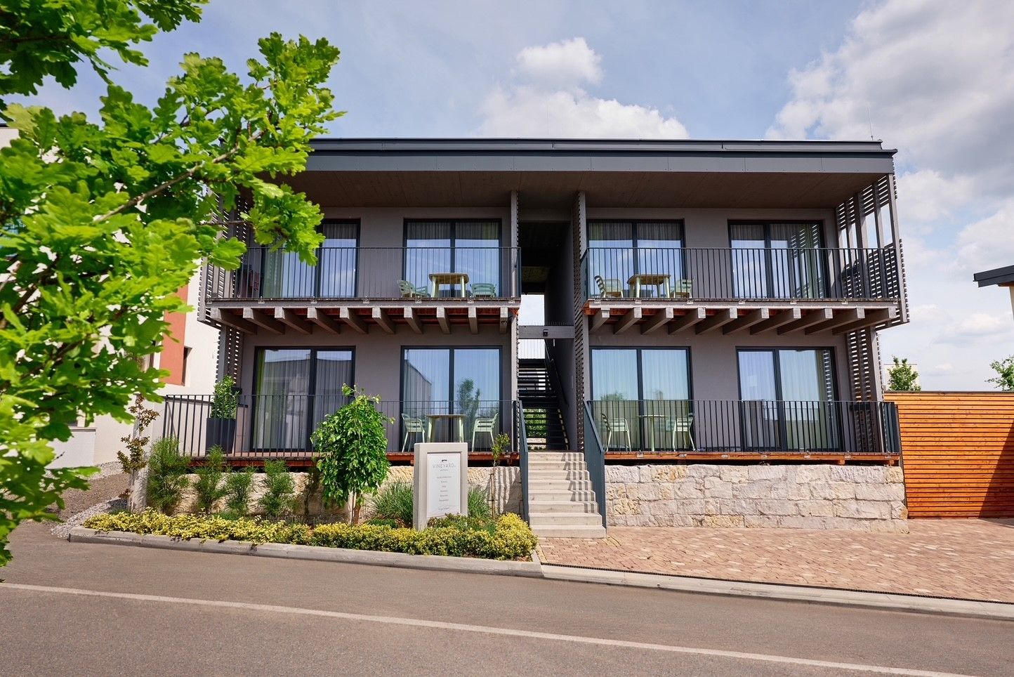 Moderne zweistöckige Apartmentanlage mit Balkonen und Treppenaufgang, umgeben von Pflanzen und Bäumen.