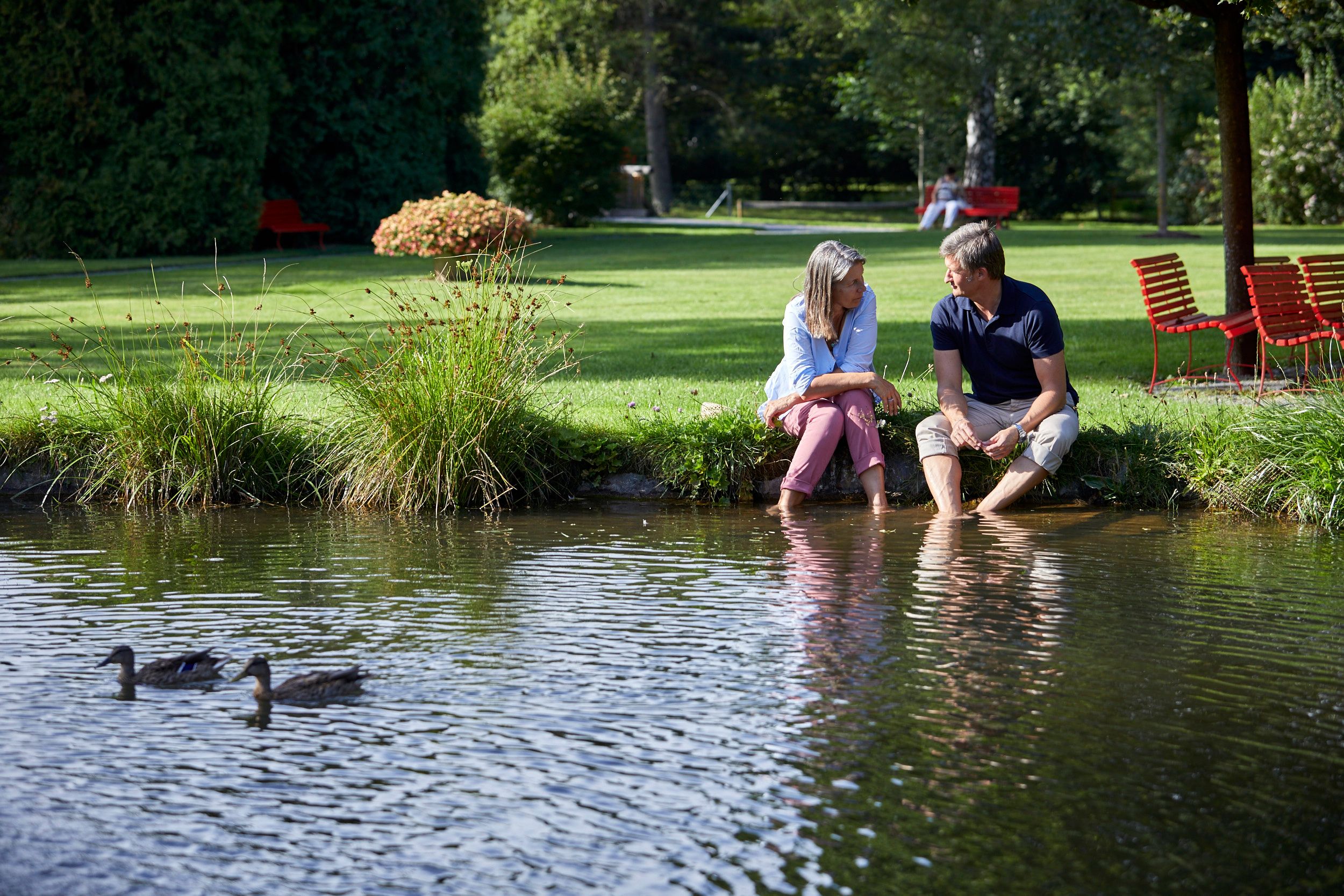 Ein Paar sitzt am Ufer eines Teichs im Kurpark Bad Schönau, die Füße im Wasser. Zwei Enten schwimmen im Vordergrund.