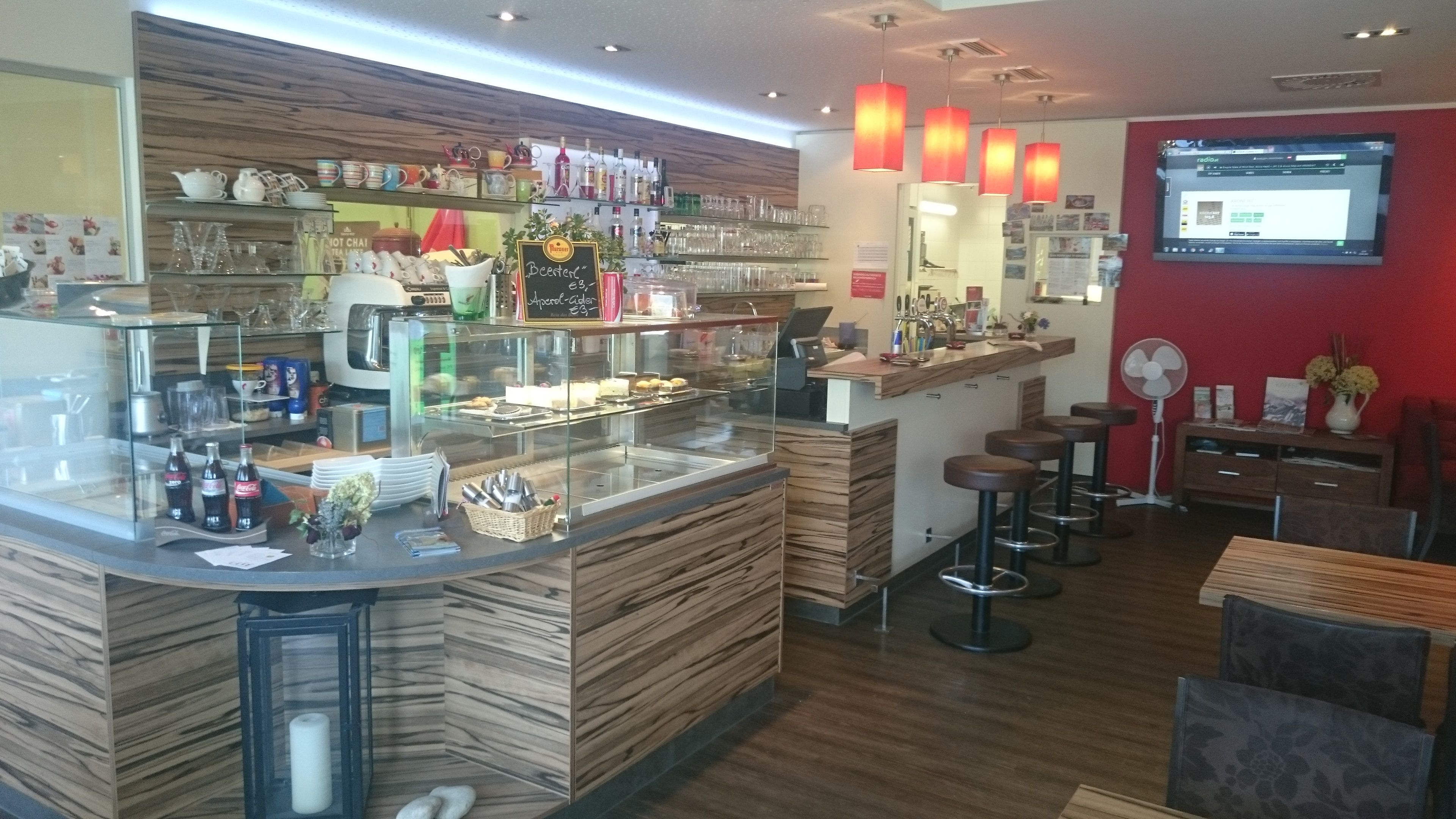 Innenansicht eines Cafés mit Theke, Barhockern und roter Wand.