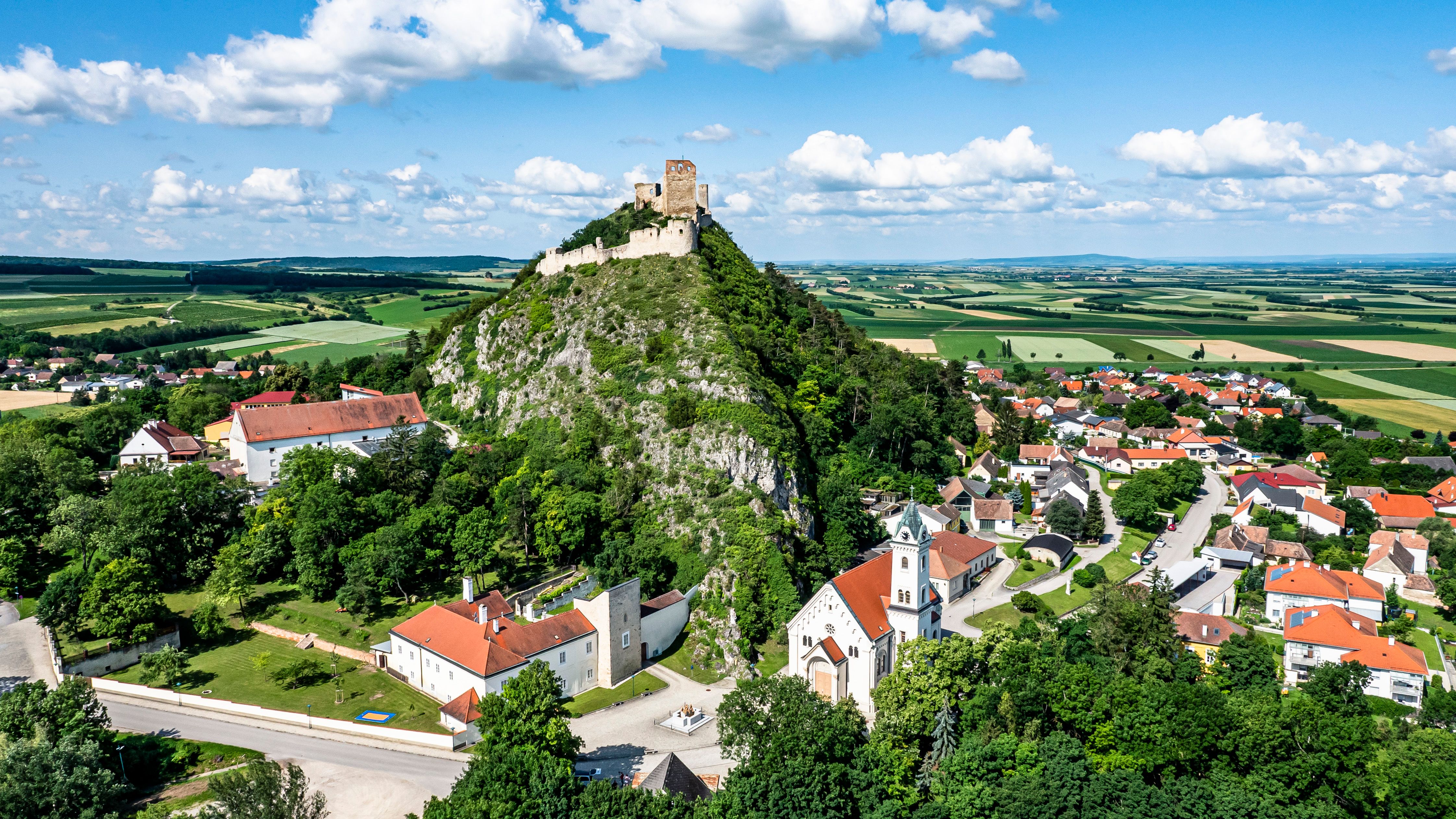 Luftaufnahme einer Burg auf einem Hügel mit umliegendem Dorf und Feldern.
