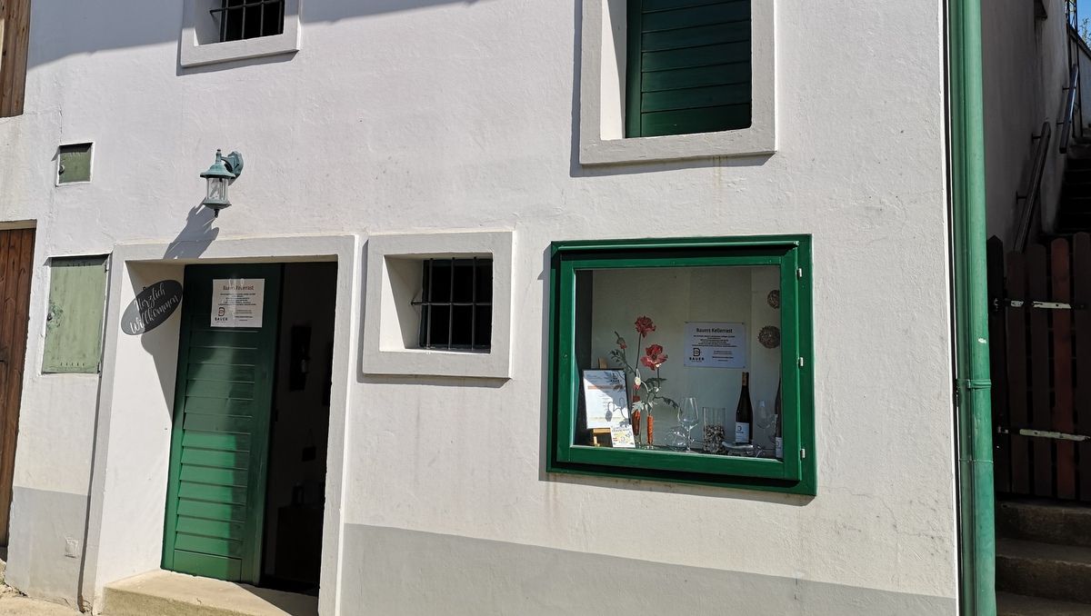 Weiße Hausfassade mit grüner Tür und Fensterrahmen. Ein Schild mit der Aufschrift 'Bauers Kellerrast' ist sichtbar.