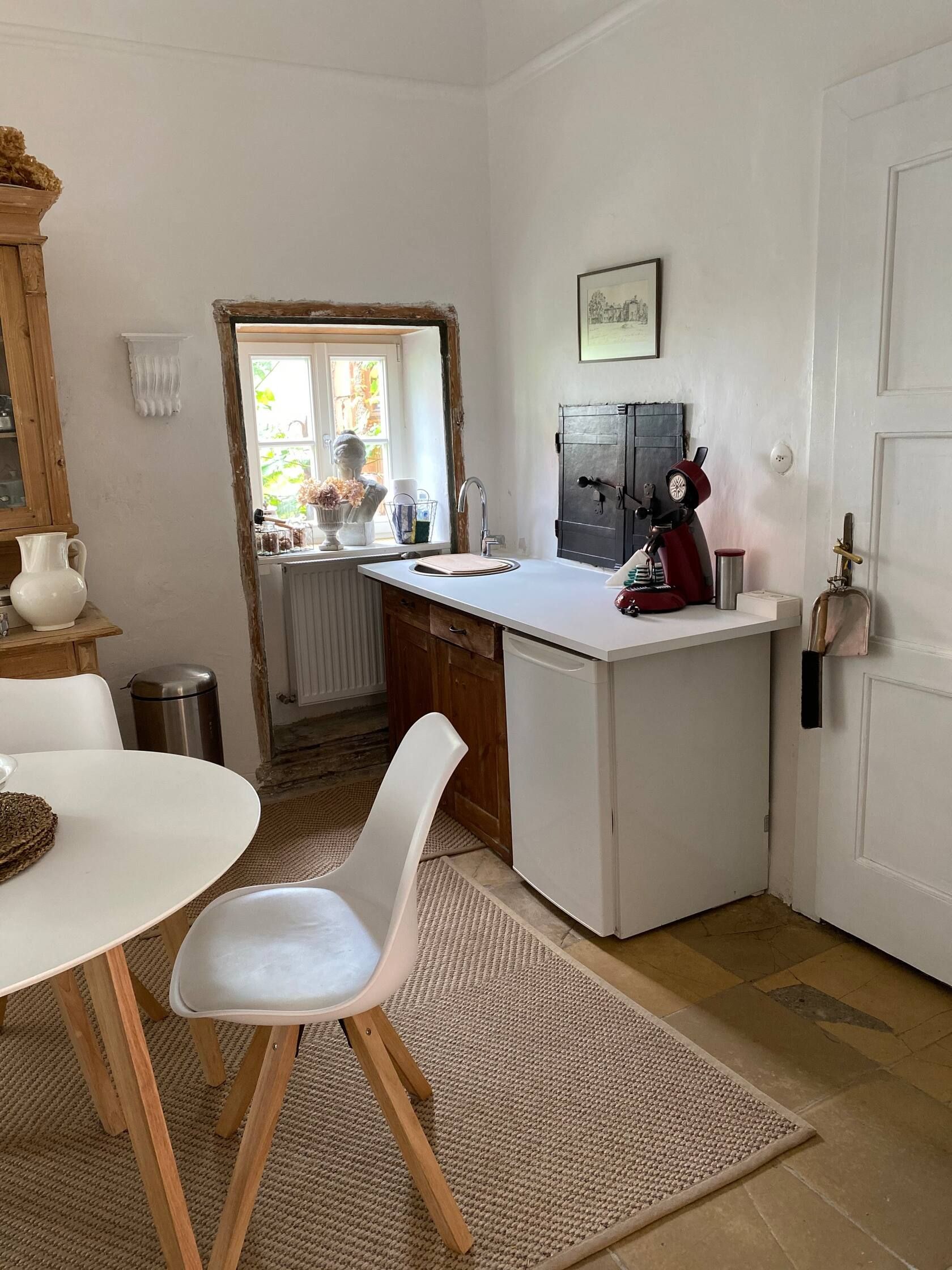 Küchenbereich mit kleinem Tisch, Stuhl, Spüle und Kaffeemaschine. Fenster mit Dekoration und helle Wände.