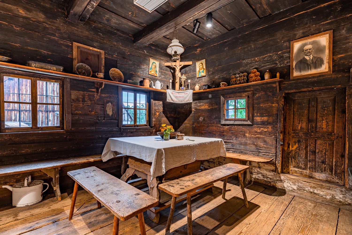 Rustikaler Raum mit Holzwänden, Tisch mit weißem Tuch, Bänken, religiösen Bildern und antiken Gegenständen.