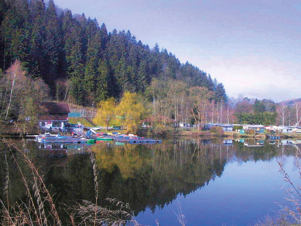 Seecampingplatz mit Wohnwagen und Booten am Ufer eines Sees, umgeben von Wald.