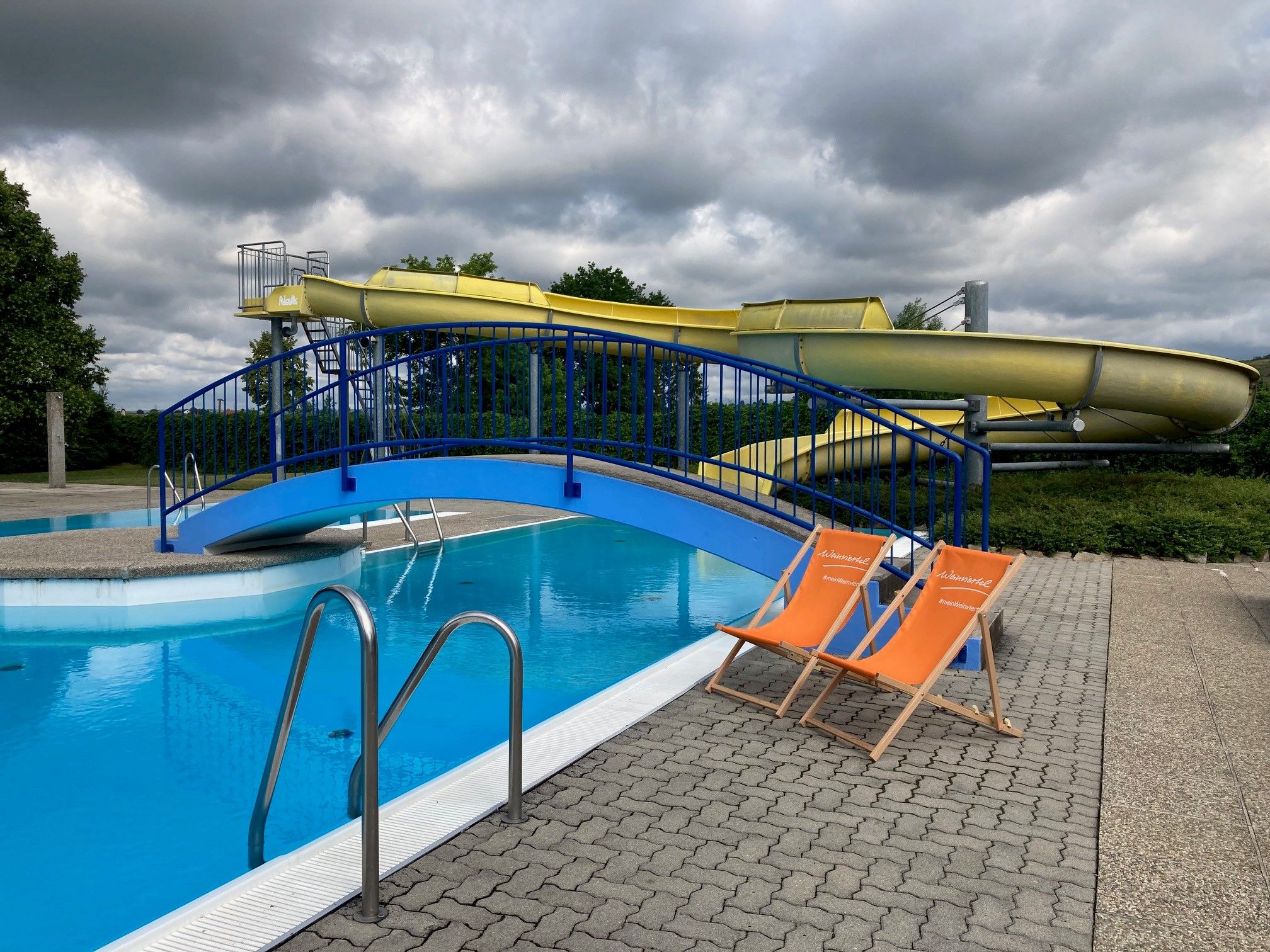 Zwei orange Liegestühle stehen neben einem Schwimmbecken mit einer blauen Brücke und einer gelben Wasserrutsche im Hintergrund.
