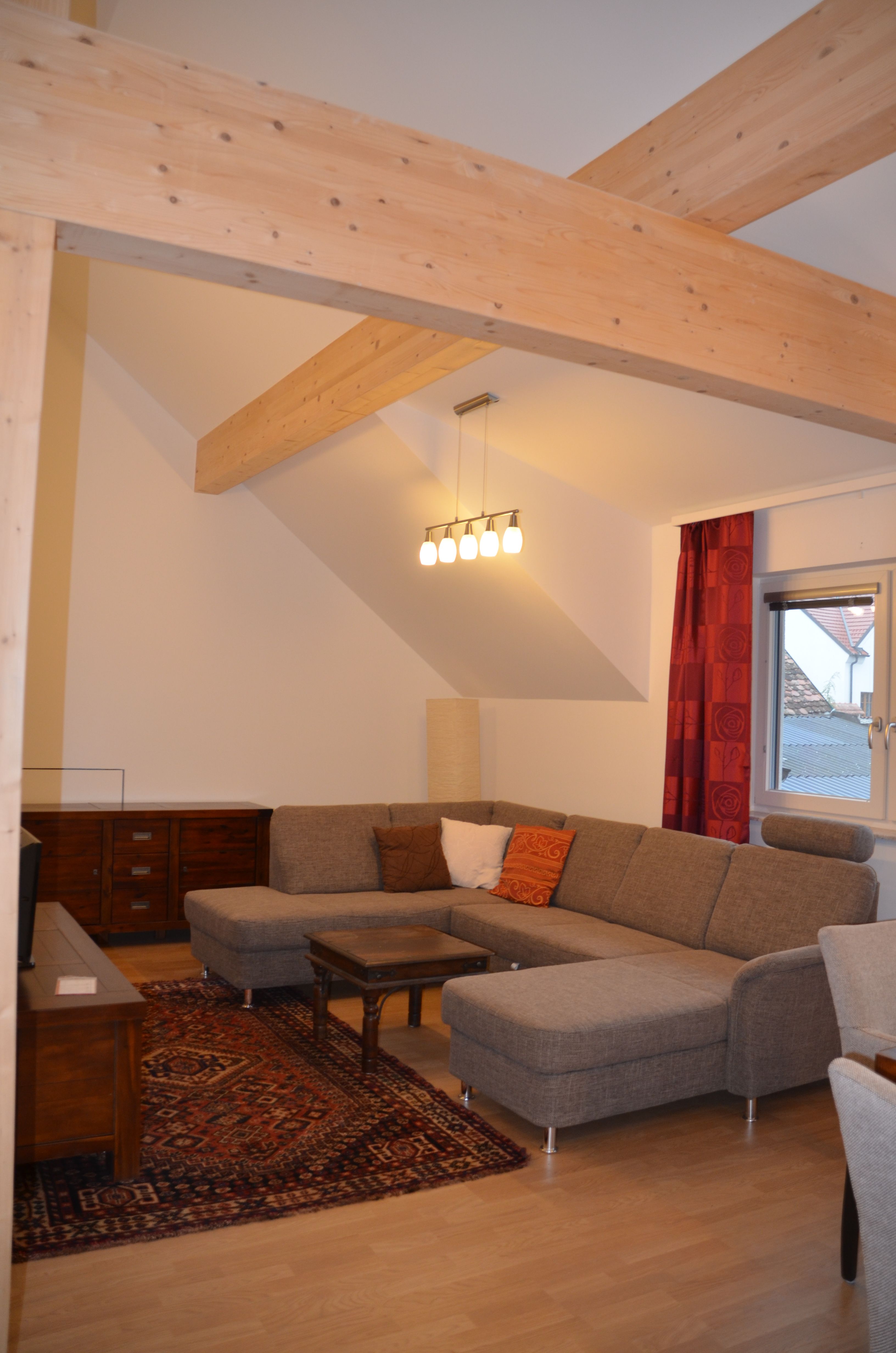 Modernes Wohnzimmer mit grauem Ecksofa, Holzbalken, Teppich und Lampe.