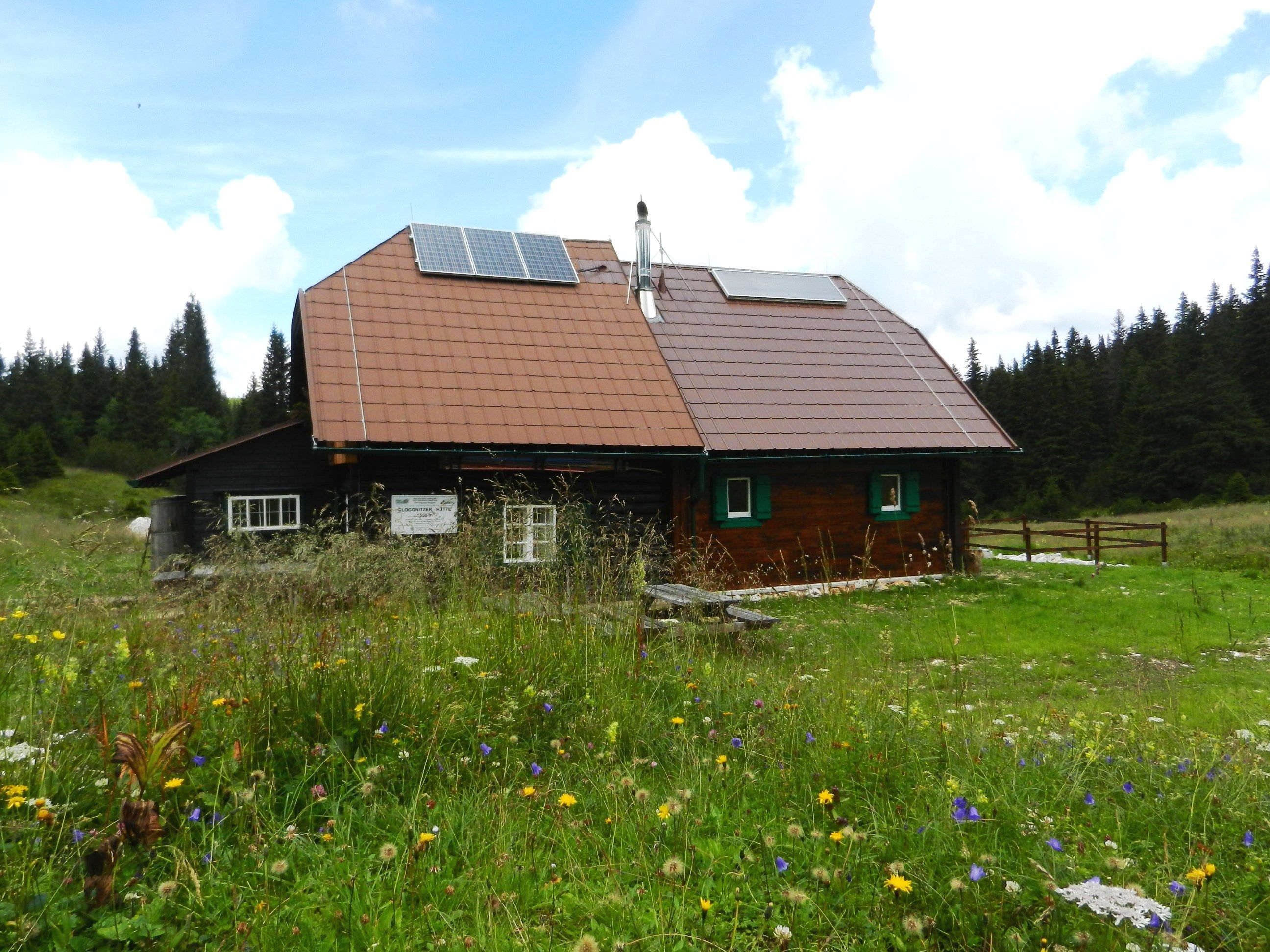 Eine Berghütte mit Solarpanelen auf dem Dach, umgeben von einer blühenden Wiese und Wald im Hintergrund.
