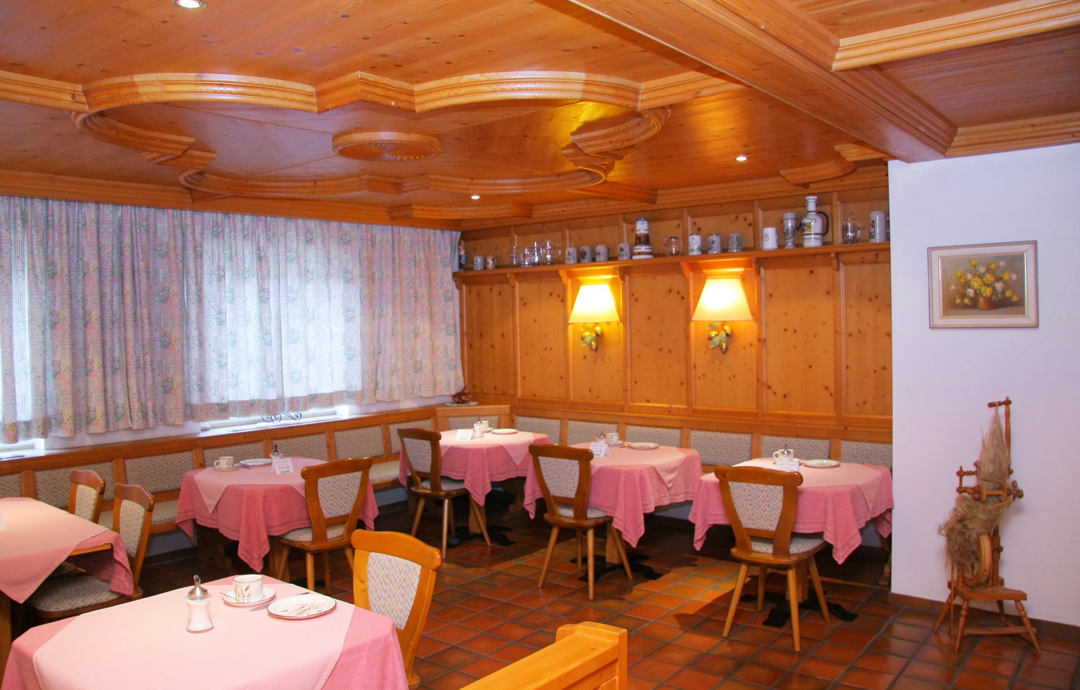 Gemütlicher Frühstücksraum mit Holzmöbeln, rosa Tischdecken und dekorativen Krügen auf einem Regal.