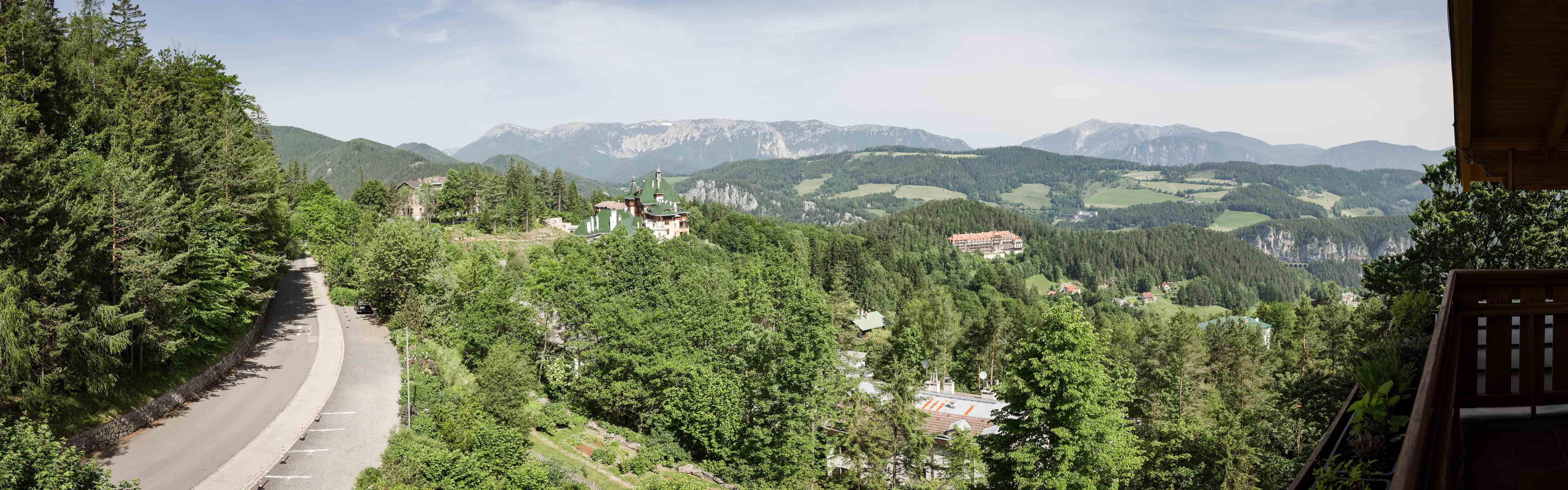 Panoramablick auf eine bergige Landschaft mit Wald, Straße und Gebäuden.