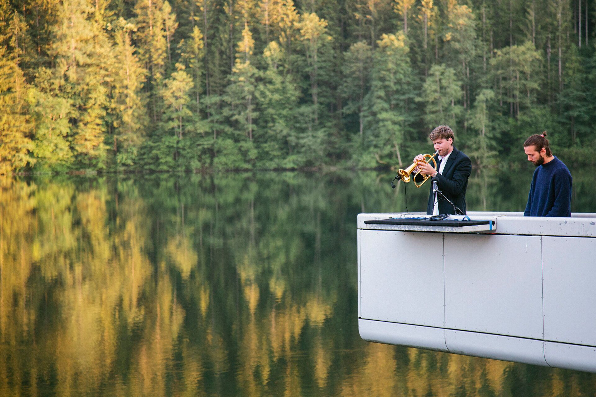 Zwei Musiker stehen auf einer schmalen, weißen Plattform über einem ruhigen See; einer spielt Trompete, der andere arbeitet an elektronischen Klängen, im Hintergrund spiegelt sich der Wald im Wasser.