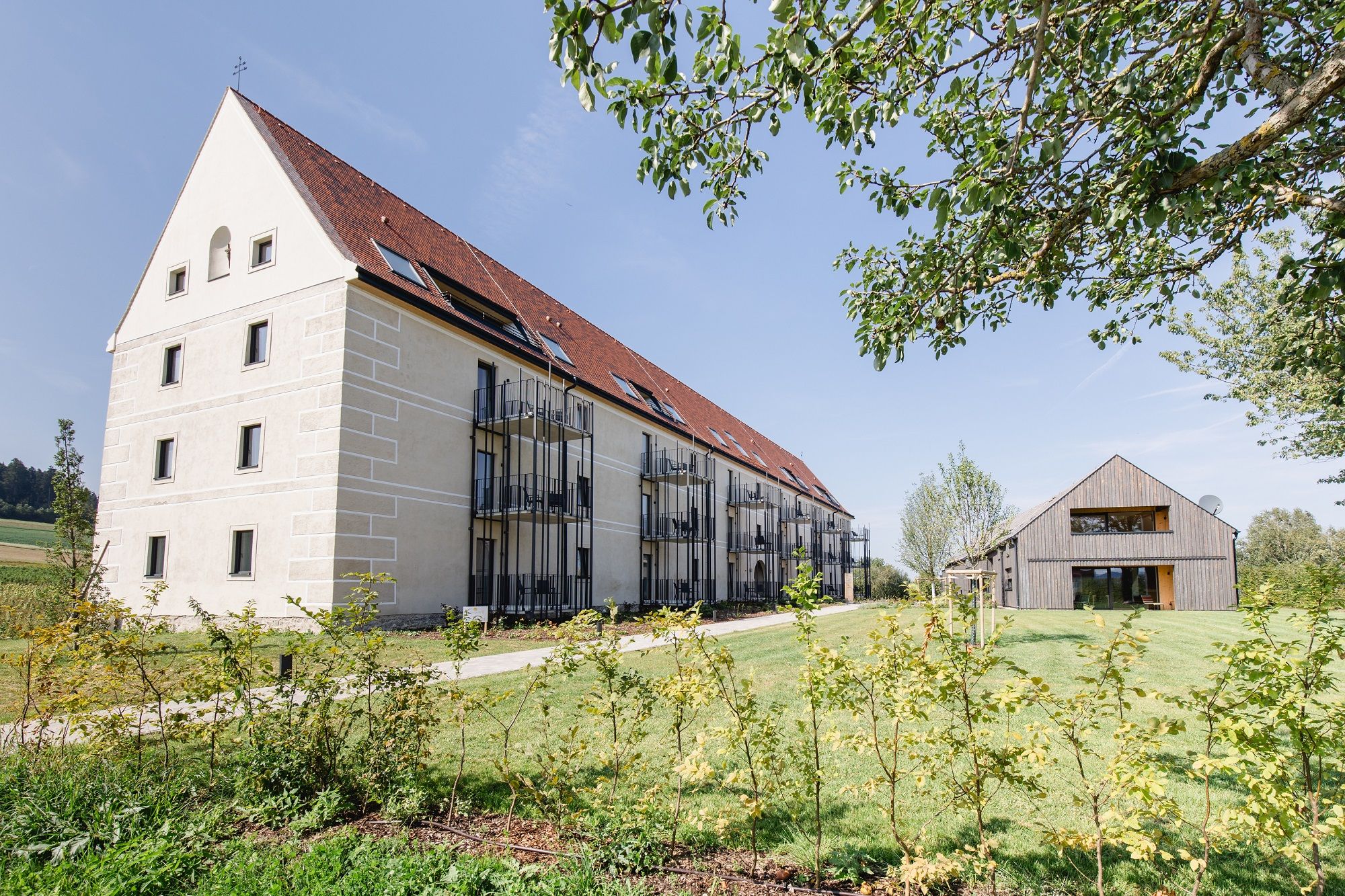 Ein großes, modernes Gebäude mit rotem Dach und Balkonen neben einem kleineren Holzhaus auf einem grünen Grundstück.