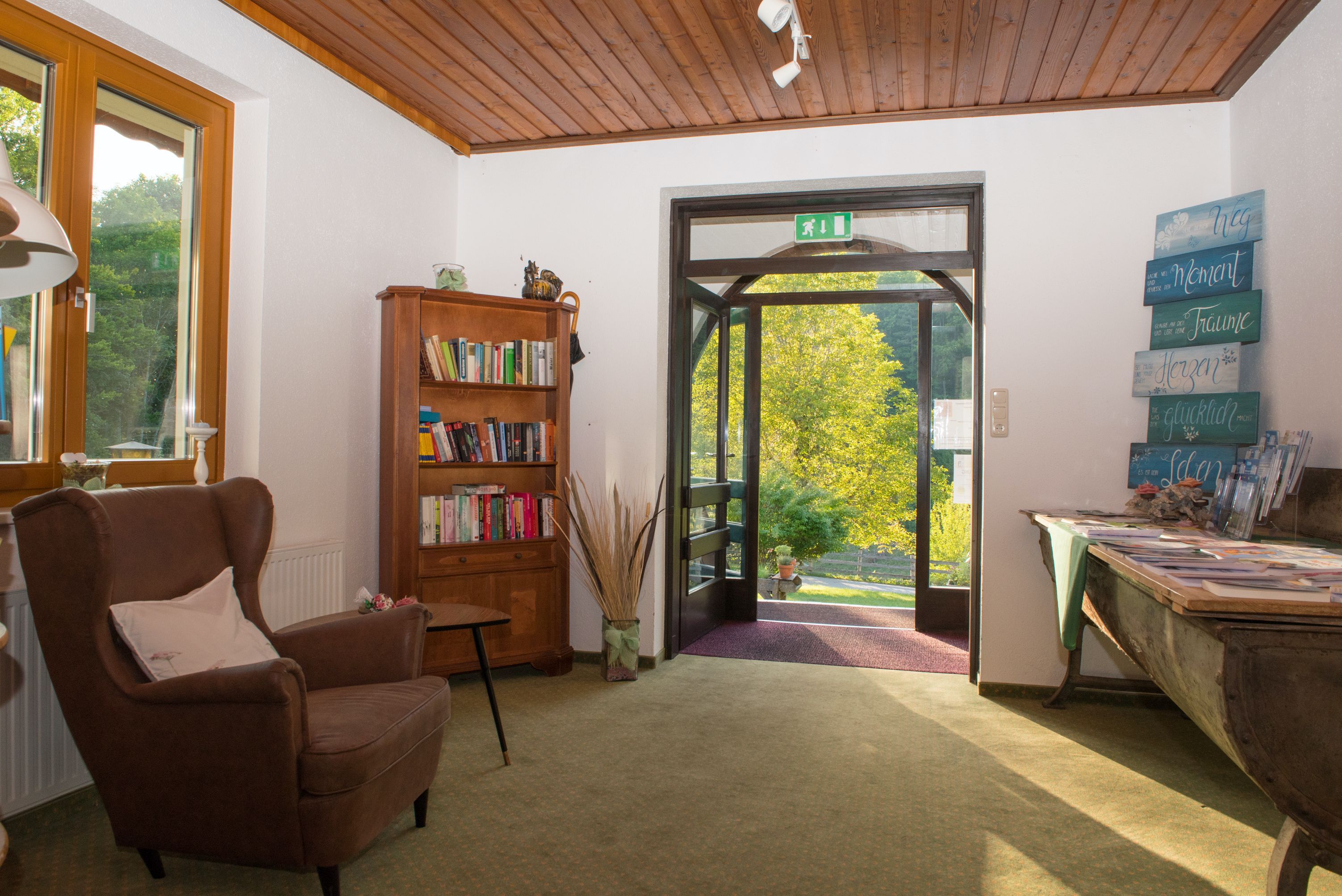 Eingangsbereich mit Sessel, Bücherregal und offenem Türbereich mit Blick ins Grüne.