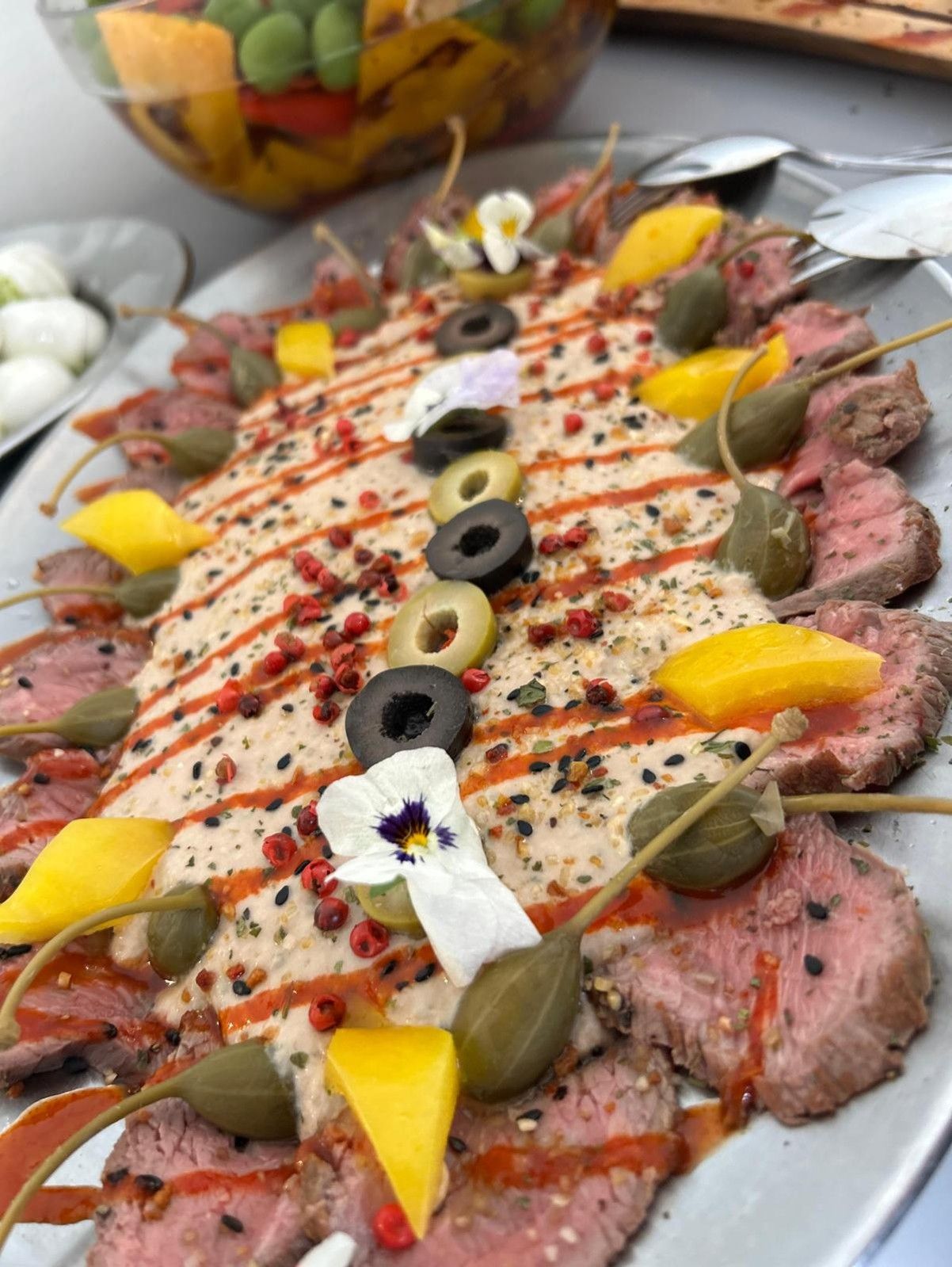 Nahaufnahme eines dekorativen Tellers mit rosa, feingeschnittenem Fleisch mit Kapern, Oliven und essbaren Blüten.