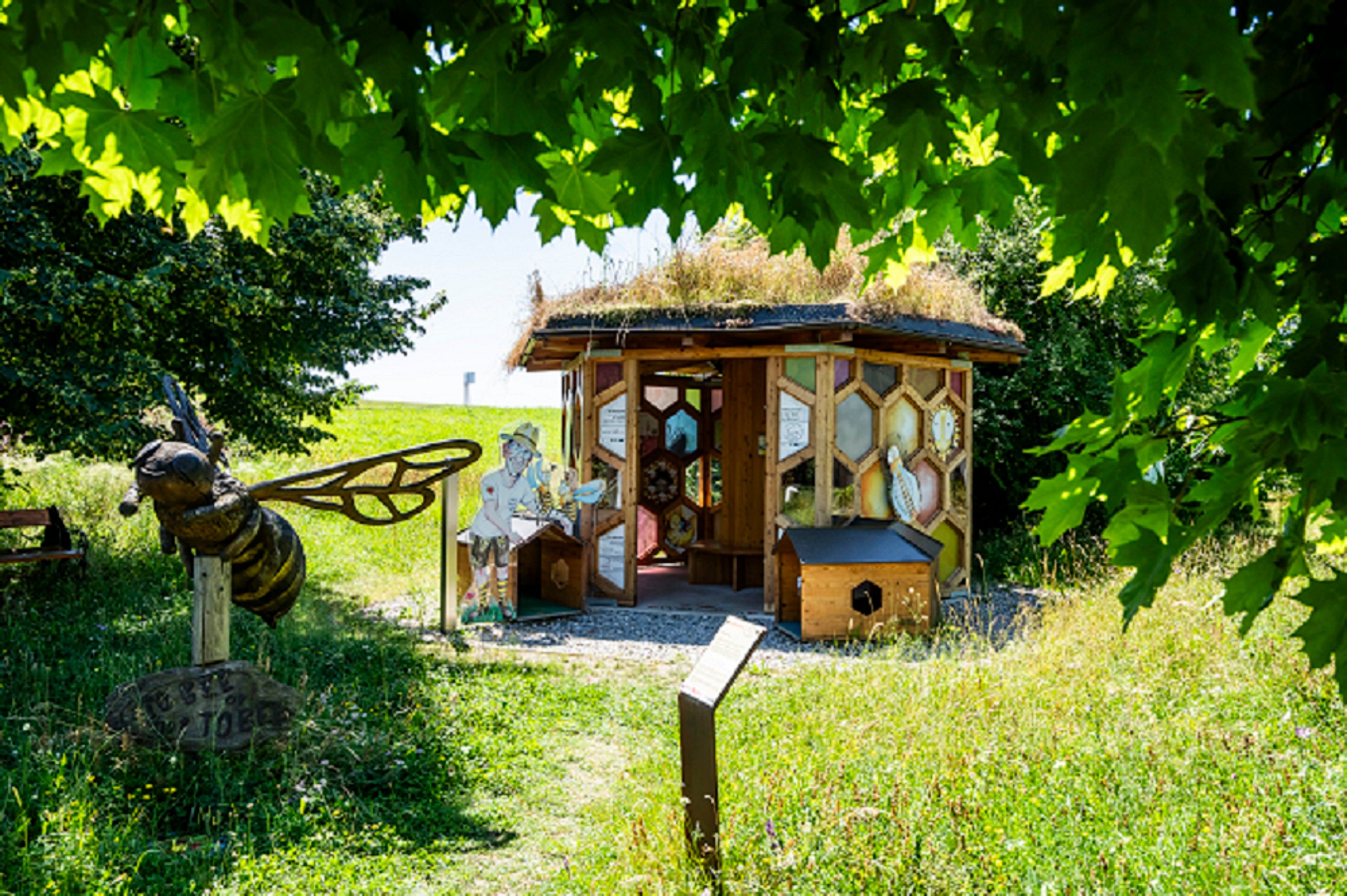 Ein kleines, hölzernes Gebäude mit Bienenmotiven und einer großen Bienenfigur im Vordergrund, umgeben von grüner Natur.