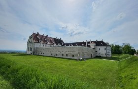 Ein gro&szlig;es, historisches Schloss mit wei&szlig;en Mauern und roten D&auml;chern steht auf einer gr&uuml;nen Wiese unter einem bew&ouml;lkten Himmel.
