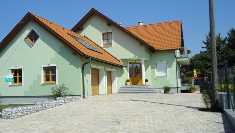 Ein modernes, zweist&ouml;ckiges Haus mit gr&uuml;nem Anstrich und roten Dachziegeln, umgeben von einem gepflasterten Hof.