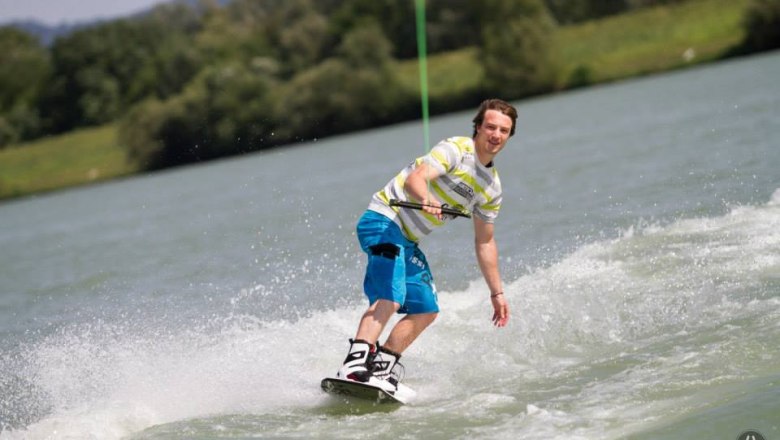 Ein Wakeboarder fährt auf einem See, umgeben von grüner Landschaft.