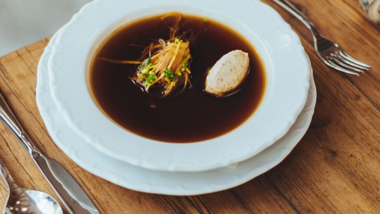 Eine klare Rindssuppe mit Gem&uuml;se und Kn&ouml;del in einem wei&szlig;en Teller auf einem Holztisch.