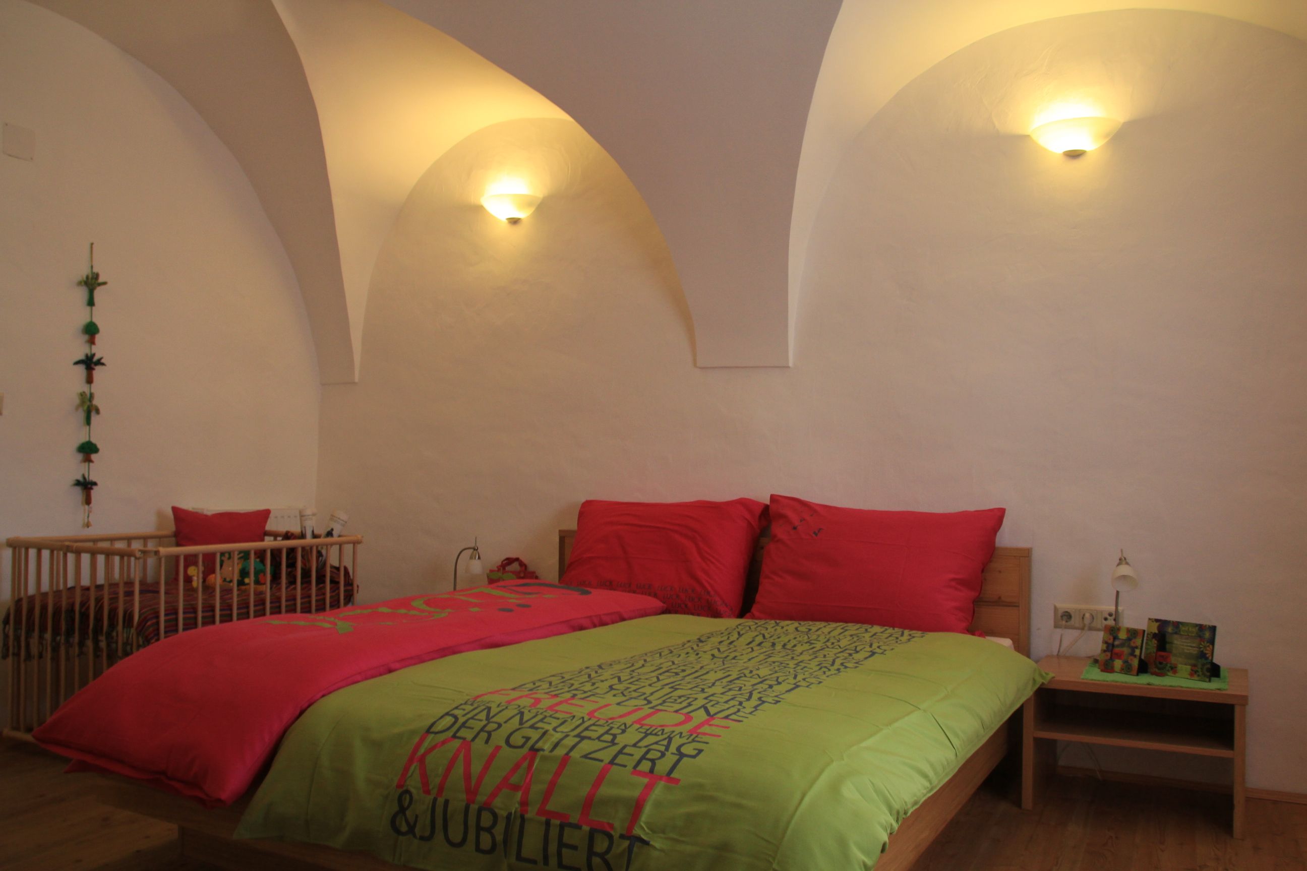 Ein gemütliches Gästezimmer mit einem Doppelbett, bunten Bettbezügen und einem Babybett im Hintergrund. Die Wände sind weiß und gewölbt.