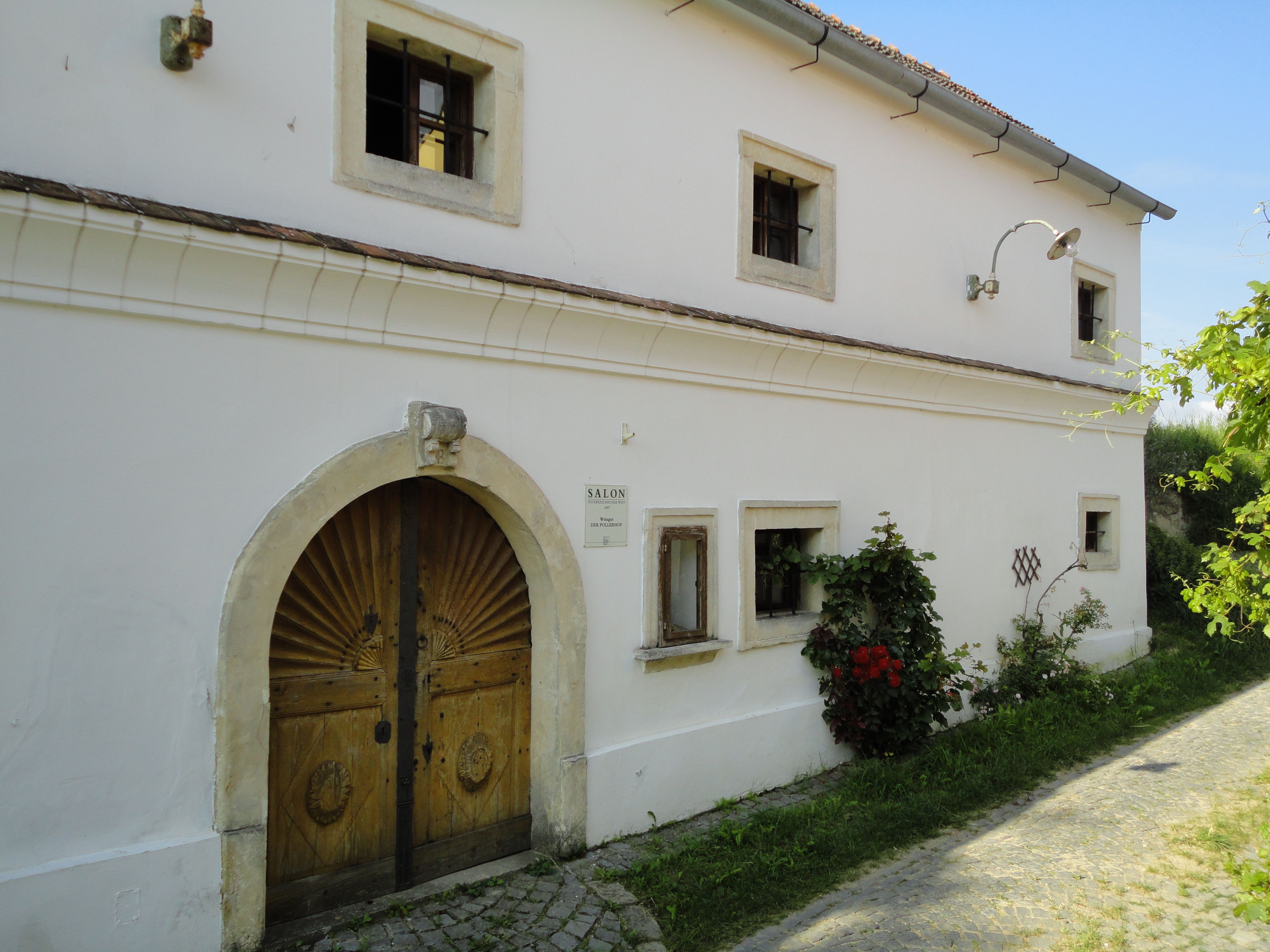 Historisches Gebäude mit Holztür und kleinen Fenstern, umgeben von Pflanzen.