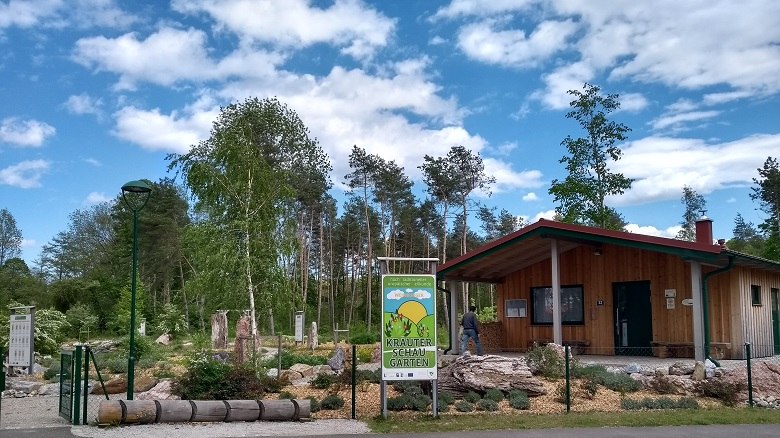 Eingang zum TEH Kr&auml;uterschaugarten mit Holzgeb&auml;ude und B&auml;umen im Hintergrund.