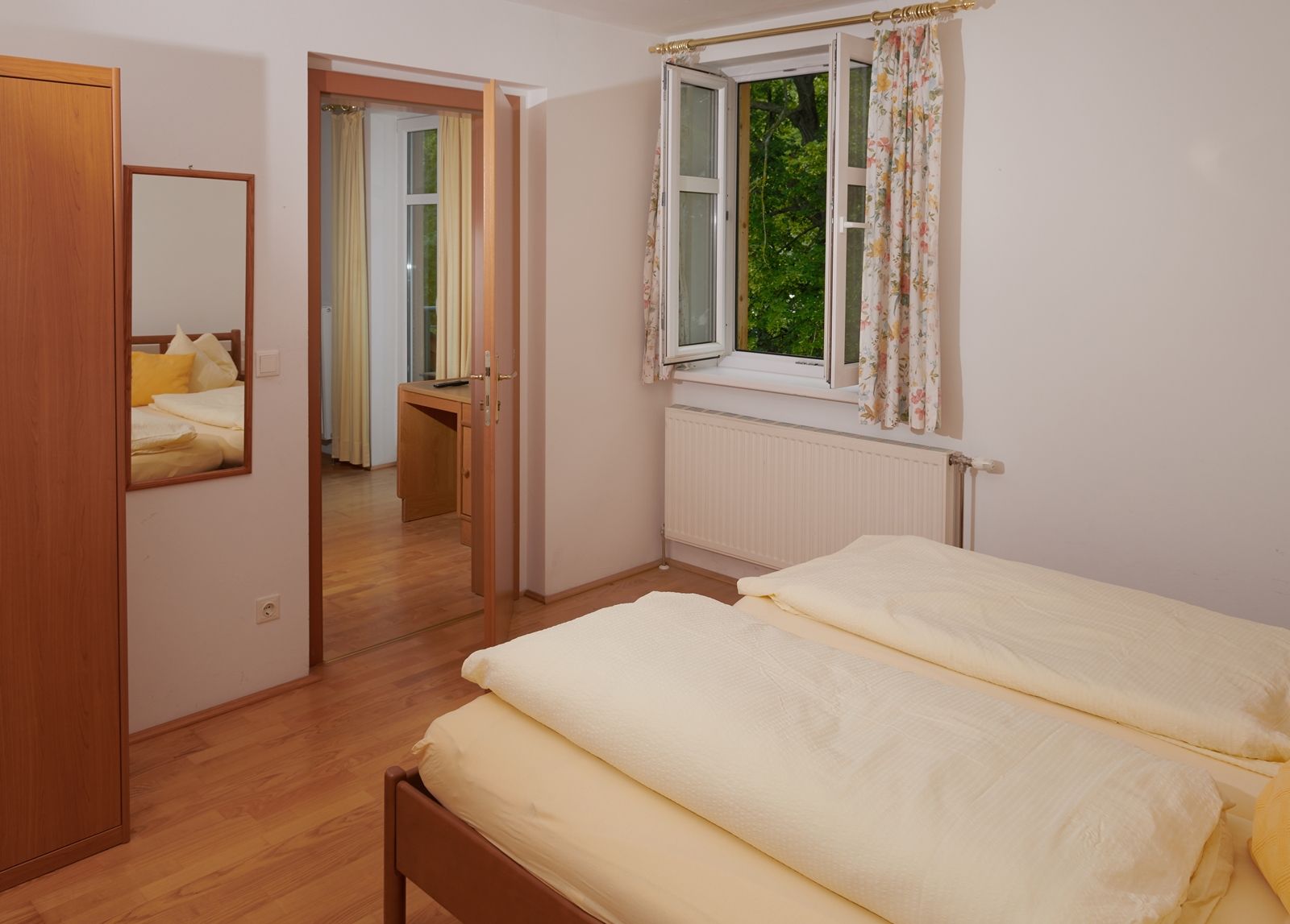 Ein Schlafzimmer mit einem Doppelbett, einem offenen Fenster mit Vorhängen und einem Spiegel an der Wand.