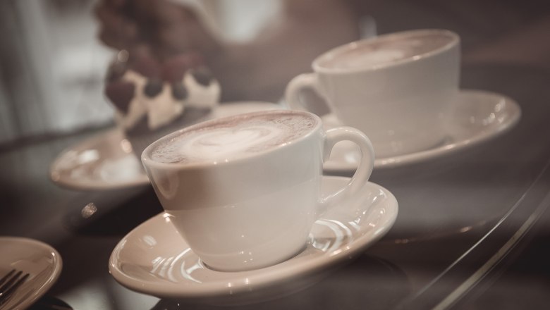 Zwei Tassen Kaffee auf Untertassen, im Hintergrund ein Dessert.