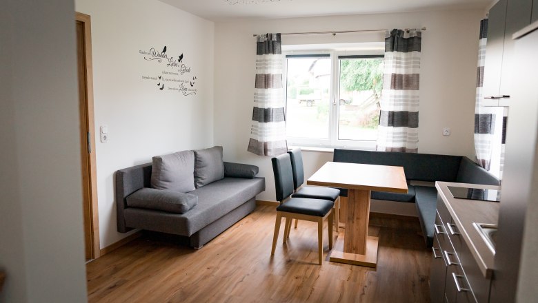 Wohn-/Esszimmer, © Manuel Hahn Moderne Küche mit grauem Sofa, Holztisch und Stühlen, Fenster mit Vorhängen.