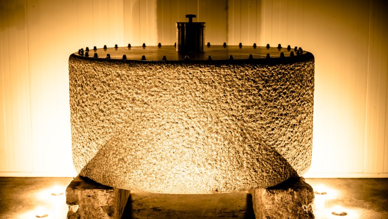 Ein gro&szlig;er, zylindrischer Pressstein aus grobem Material, beleuchtet von warmem Licht, steht auf zwei Steinen in einem Raum.