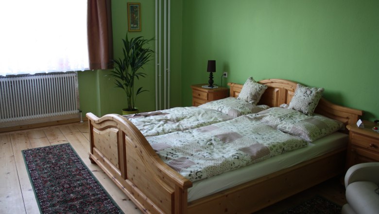 Gästezimmer, © Majewsky Zant Ein gemütliches Gästezimmer mit grün gestrichenen Wänden, einem Holzbett, Nachttischen, einer Pflanze und einem Teppich auf Holzboden.