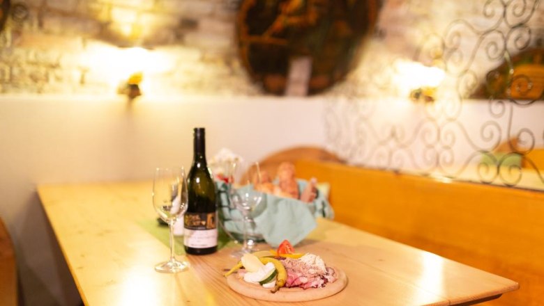 Ein Tisch in einem rustikalen Raum mit Weinflasche, Gl&auml;sern und einer Jause mit Brot und Aufschnitt.