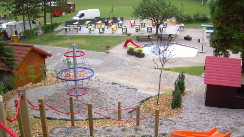 Spielplatz mit Klettergerüst, Rutsche und kleinem Pool, umgeben von Bäumen und Wiese.