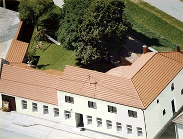 Luftaufnahme eines Geb&auml;udekomplexes mit rotem Dach und Innenhof mit B&auml;umen und Spielplatz.