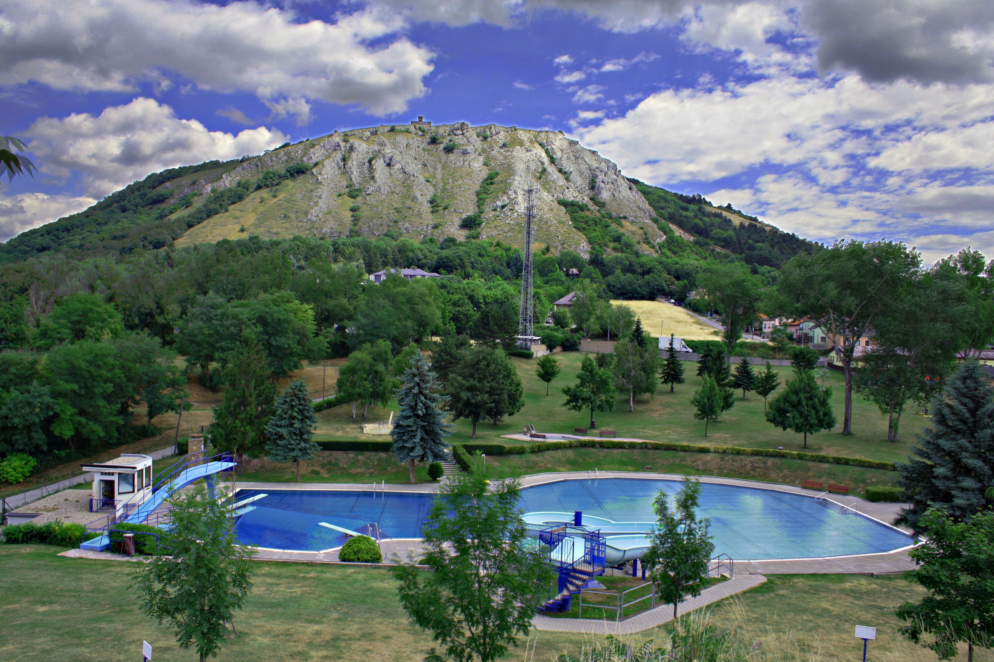Ein Freibad mit einem runden Becken und einer Rutsche, umgeben von Bäumen, vor einem bewaldeten Hügel mit einer felsigen Spitze.