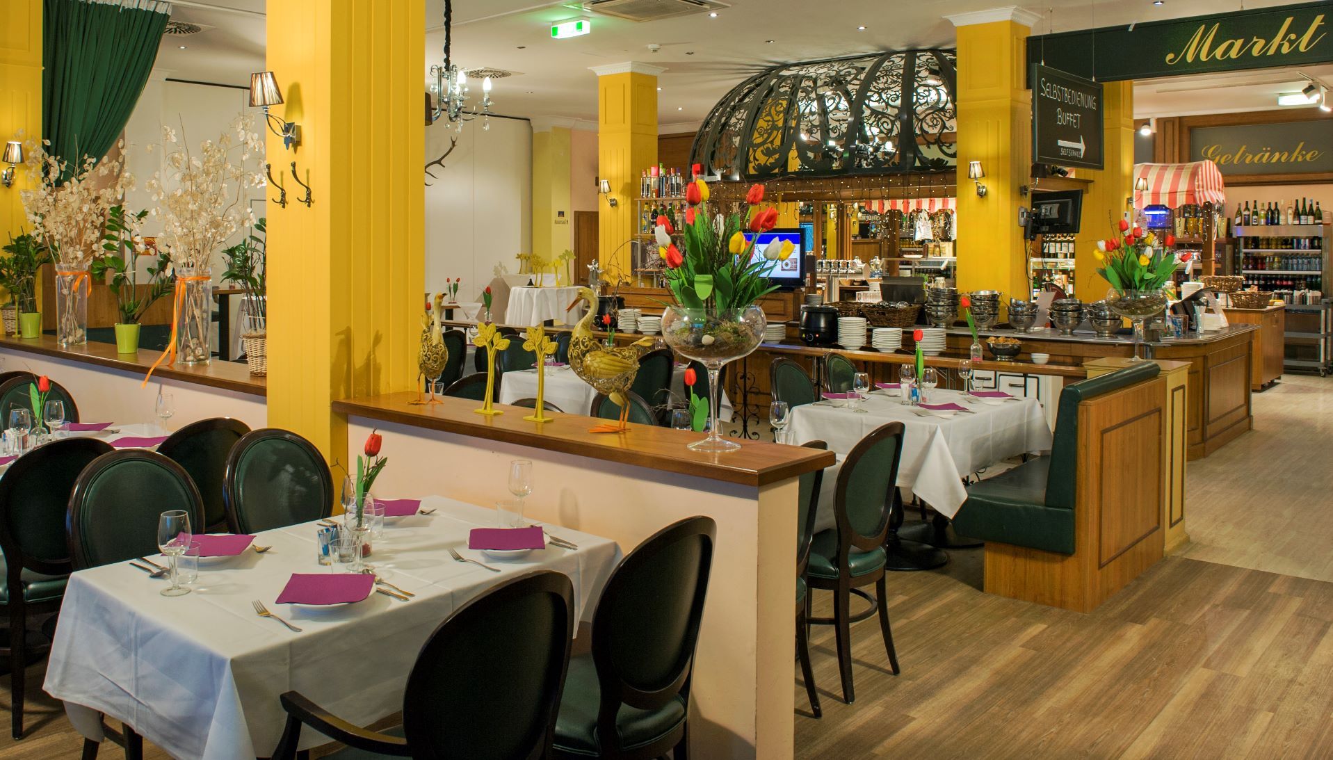 Restaurant mit Tischen, welche von Halbwänden und gelben Betonsäulen getrennt werden. Im Hintergrund sieht man dezent die Küche.