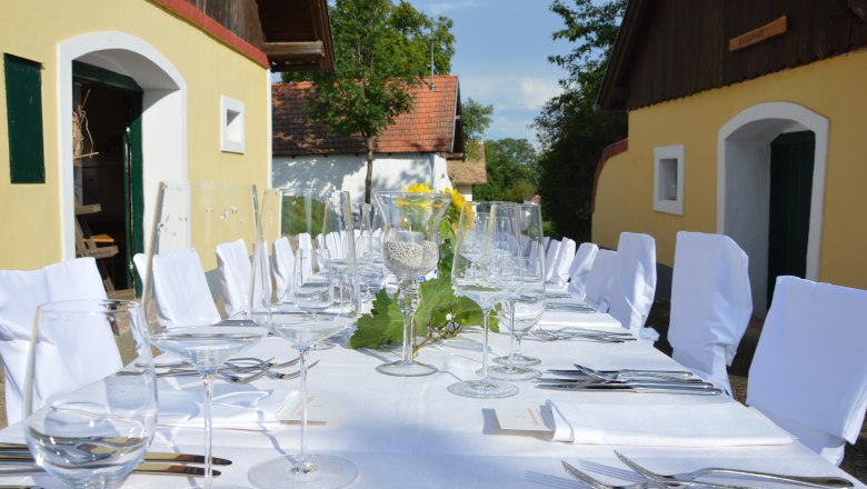 Langer, festlich gedeckter Tisch im Freien mit wei&szlig;en Tischdecken und St&uuml;hlen, umgeben von gelben Geb&auml;uden.