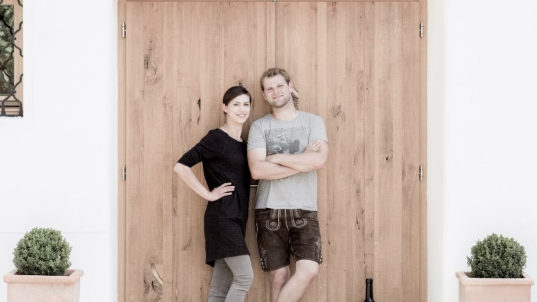 Zwei Personen stehen vor einer Holzt&uuml;r, daneben eine gro&szlig;e Weinflasche und Pflanzen in T&ouml;pfen.