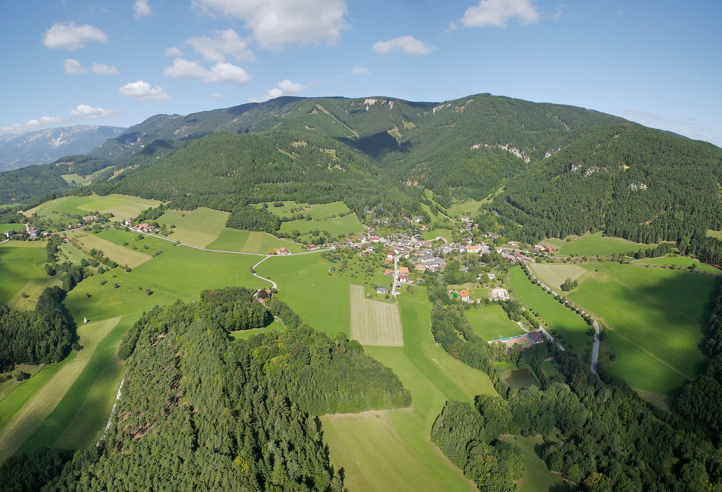 Luftaufnahme der Gemeinde Prigglitz mit grünen Feldern und Wäldern, umgeben von Hügeln.
