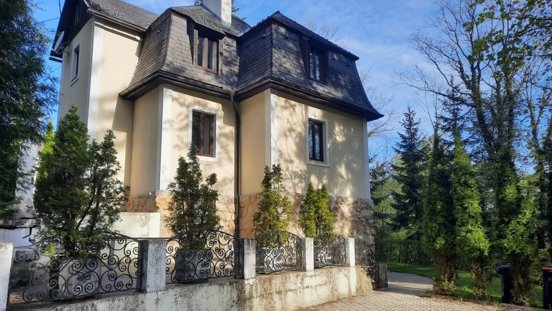 Villa Agnes, © Dr. Eva Dirnbacher Eine zweistöckige Villa mit Mansardendach, umgeben von Bäumen und einem schmiedeeisernen Zaun.