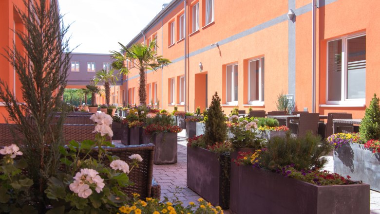 winrooms_gastgarten, © Winrroms Betriebs GmbH Ein sonniger Gastgarten mit orangen Gebäuden, Palmen und bunten Blumen in großen Pflanzkübeln.
