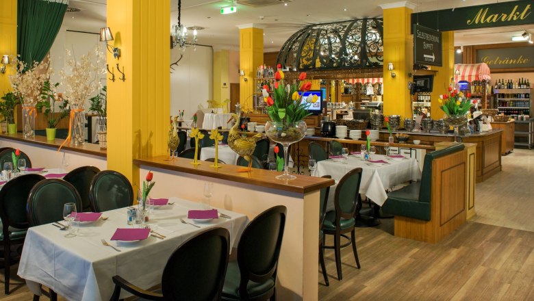 Restaurant mit Tischen, welche von Halbwänden und gelben Betonsäulen getrennt werden. Im Hintergrund sieht man dezent die Küche.