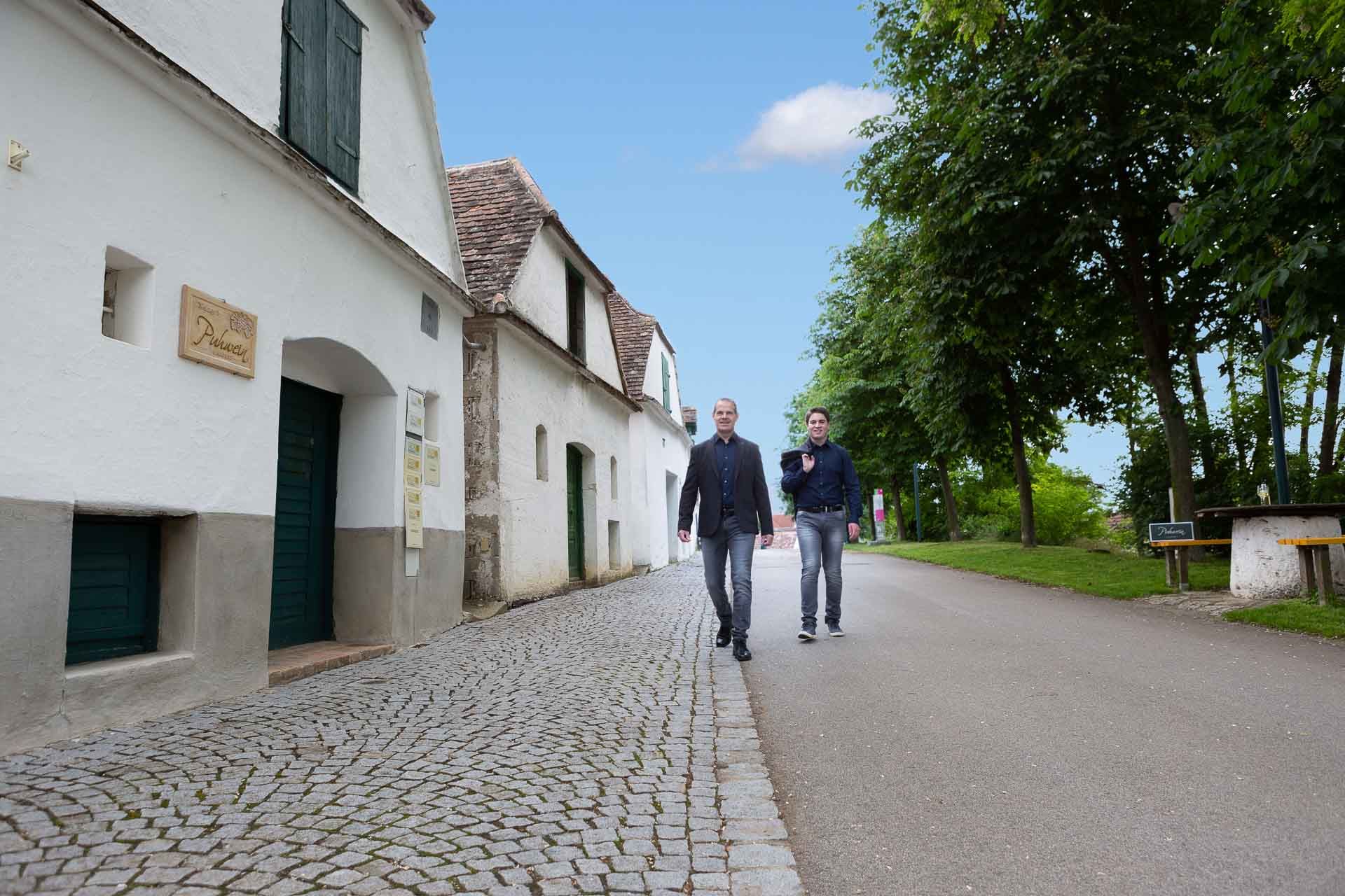 Zwei Männer gehen auf einer gepflasterten Straße entlang, vorbei an weißen Gebäuden und Bäumen.