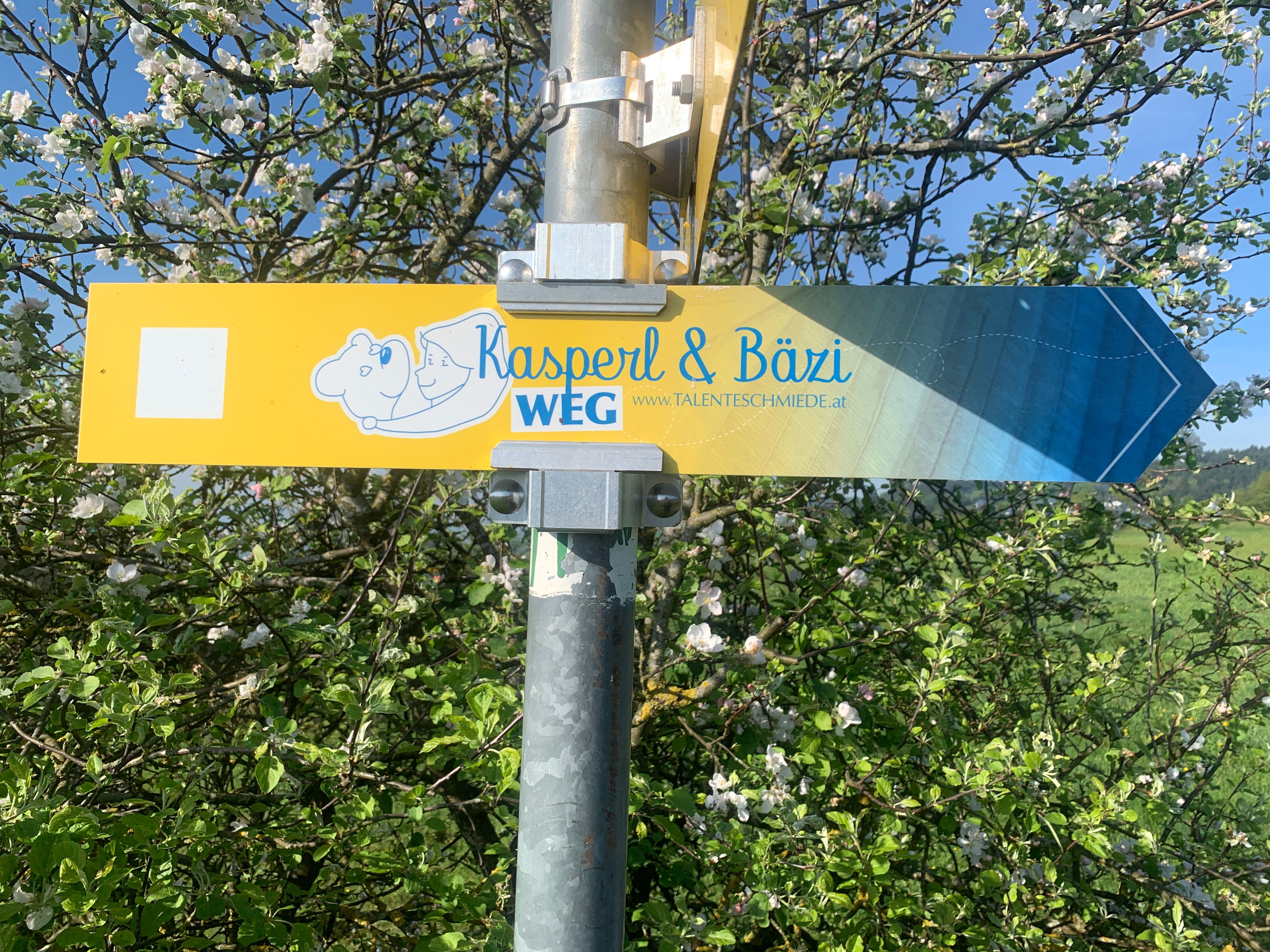 Wegweiser mit der Aufschrift 'Kasperl & Bäzi WEG' vor blühendem Baum.