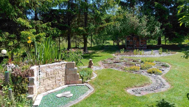 Ein gepflegter Garten mit Steinen, Pflanzen und einem Insektenhotel im Hintergrund.